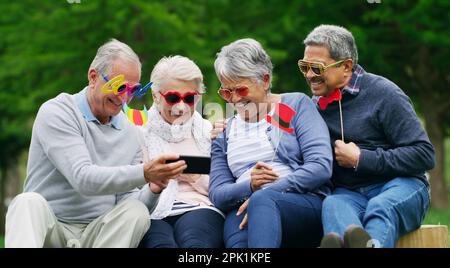 Sachez vous amuser et vous ne serez jamais vieux. un groupe d'hommes et de femmes âgés heureux prenant des selfies tout en portant des lunettes amusantes au parc. Banque D'Images