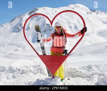 Une femme skieuse pose à l'intérieur d'un cadre en forme de coeur à la station de ski de Tignes - Val d'Isère, en France, avec tous les skis de montagne et les tenues rouges, par temps ensoleillé. Touris Banque D'Images