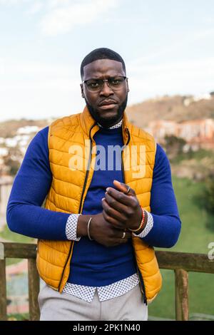 Jeunes afro-américains élégants qui se posent sur les balustrades du parc avec vue sur la ville, enfilant un pull bleu décontracté, une veste jaune et des lunettes. Banque D'Images