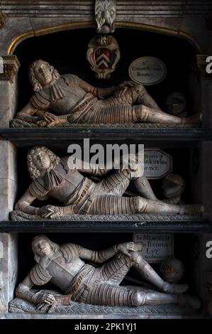 Les patriarches du XVIIe siècle de la riche famille Fettiplace, portant une armure dorée caroléenne, se reposent détendus, tendus sur leurs coudes, sur des étagères à plusieurs niveaux dans l'église paroissiale de St Mary à Swinbrook, dans la vallée du Windrush, dans l'Oxfordshire, en Angleterre, au Royaume-Uni. Le somptueux monument baroque de 1686 contraste avec un prédécesseur de 1613 qui abrite trois anciens hommes Fettiplace, qui étaient rigidement dans une armure Tudor non ornée. Banque D'Images