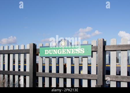 Un panneau pour la gare s'arrête sur une clôture à la gare de Dungeness sur le Romney, Hythe et Dymchurch Light Railway à Dungeness, Kent. Avec un train Banque D'Images