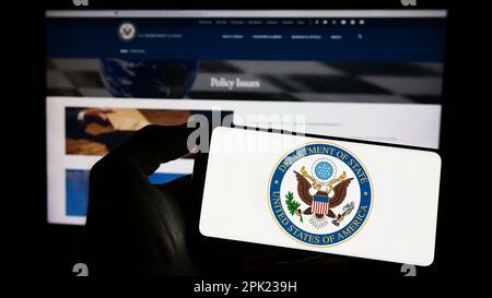 Personne tenant un téléphone portable avec le sceau du Département d'État des États-Unis (DOS) sur l'écran en face de la page Web. Mise au point sur l'affichage du téléphone. Banque D'Images
