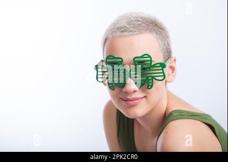 Portrait d'une jeune femme caucasienne avec une coupe de cheveux d'un homme dans un t-shirt vert et des lunettes gaies sur un fond blanc. La jeune fille célèbre St patrick Banque D'Images