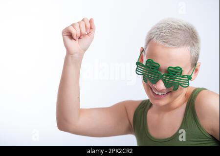 Portrait d'une jeune femme caucasienne avec une coupe de cheveux d'un homme dans un t-shirt vert et des lunettes gaies sur un fond blanc. La jeune fille célèbre St patrick Banque D'Images