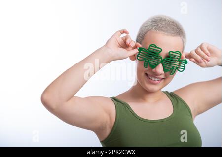 Portrait d'une jeune femme caucasienne avec une coupe de cheveux d'un homme dans un t-shirt vert et des lunettes gaies sur un fond blanc. La jeune fille célèbre St patrick Banque D'Images