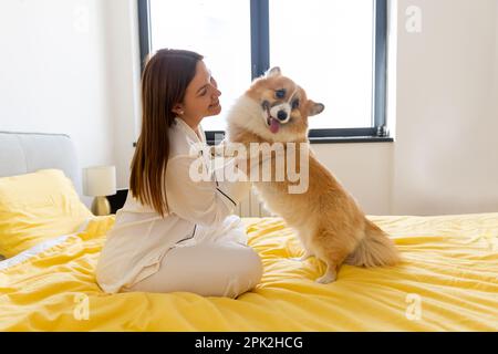 une jeune fille avec un chien corgi joue sur le lit et une journée lumineuse dans la chambre, la vie avec un chien, un ami d'une personne Banque D'Images