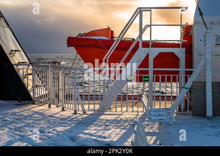 Partie d'un canot de sauvetage orange sur un pont de ferry couvert de neige et de glace, ciel spectaculaire et ensoleillé au coucher du soleil. De Moskenes à Bodø, Lofoten, Norvège. Banque D'Images