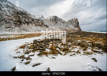 Petite route et grande montagne rocheuse dans un paysage côtier enneigé, beauté sauvage sur la plage Unstad, Lofoten, Norvège. Banque D'Images