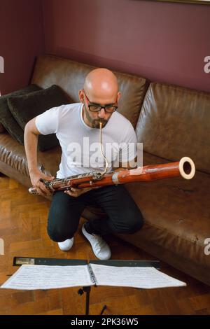 vue verticale en haut du jeune latino-américain, chauve dans des lunettes, jouant le basson étudiant la musique lecture feuille de musique assis sur un canapé dans le salon de Banque D'Images