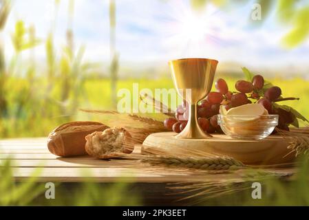 Célébration eucharistique avec coupe de calice et hôtes consacrés avec du pain et des raisins sur table en bois dans la campagne. Vue avant. Banque D'Images