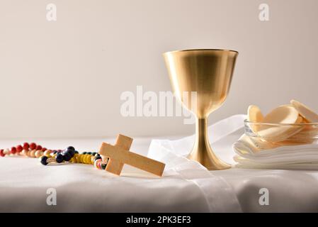 Coupe de calice au vin et hôtes consacrés dans un récipient sur table avec nappe et croix chrétienne en bois et fond blanc isolé. Vue avant Banque D'Images