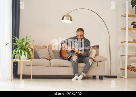 Pratique de l'écriture de chansons. Homme dans des vêtements décontractés et avec guitare acoustique est à l'intérieur Banque D'Images