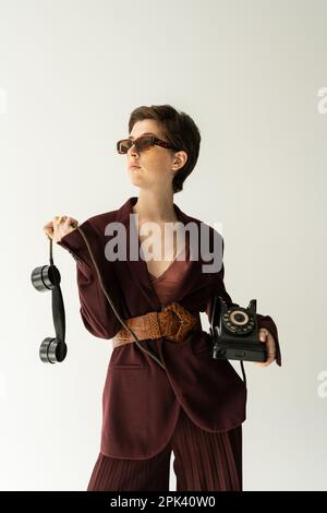 brunette femme en lunettes de soleil et blouson brun bordeaux avec ceinture en cuir posant avec téléphone vintage isolé sur gris, image de stock Banque D'Images
