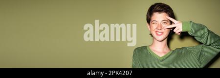 une femme brune souriante se tord à l'appareil photo et montrant le signe de paix près du visage sur fond vert, bannière, image de stock Banque D'Images