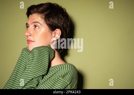 jeune et rêveuse femme dans le pull à rayures tendance regardant loin sur fond vert, image de stock Banque D'Images
