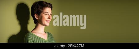 femme brunette insouciante en pull rayé regardant loin sur fond vert avec ombre, bannière, image de stock Banque D'Images