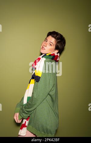 jeune et positive femme en pull-over tendance et foulard multicolore à rayures regardant l'appareil photo sur fond vert, image de stock Banque D'Images