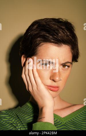 portrait de la femme fauchée avec de courts cheveux de brunette touchant le visage et regardant l'appareil photo sur fond vert, image de stock Banque D'Images