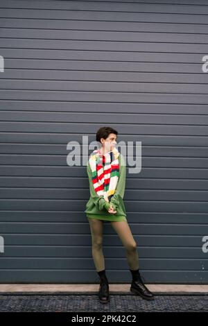 pleine longueur de femme tendance en pull-over et foulard rayé regardant loin près du mur gris sur la rue de ville, image de stock Banque D'Images