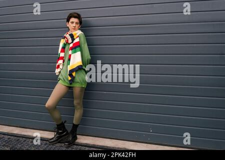 pleine longueur de femme en foulard rayé et bottes en cuir noir posant près du mur gris sur la rue, image de stock Banque D'Images