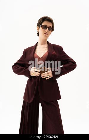 modèle brunette en combinaison marron bordeaux et lunettes de soleil touchant la ceinture en cuir isolée sur blanc, image de stock Banque D'Images