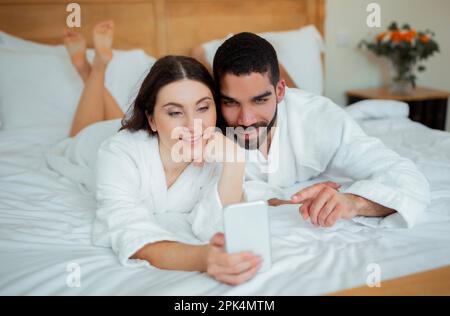 Divers conjoints utilisant un téléphone portable naviguer sur Internet dans le lit à l'intérieur Banque D'Images
