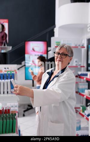 Pharmacien senior femme debout à côté des étagères de pharmacie arranger des paquets de pilules pour aider les clients, travailleur offrant des services médicaux et de soutien. Diverses personnes en pharmacie achetant des vitamines Banque D'Images