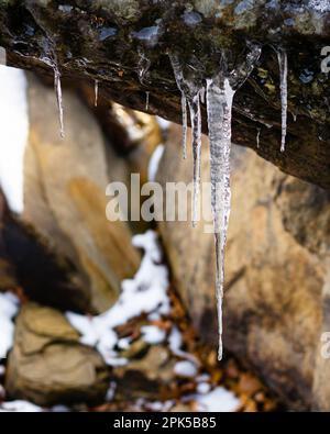 Détail gros plan d'une grande glace avec de petites glaces suspendues à une formation de roche avec des roches chaudes et de la neige en arrière-plan Banque D'Images