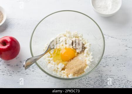 Bol en verre avec fromage cottage, œuf, cassonade, sucre vanillé entouré de pomme, farine de riz sur fond texturé gris clair, vue du dessus. Cuisine Banque D'Images