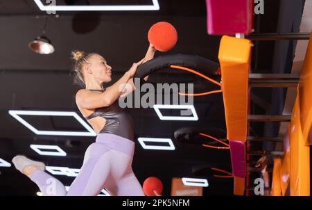 Femme européenne active dans des vêtements de sport jouant au basket-ball tout en rebondissant sur des trampolines colorés à l'intérieur dans le centre de sports de loisirs Banque D'Images