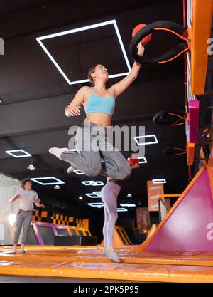 Une jeune femme européenne gaie dans des vêtements de sport jouant au basket-ball tout en rebondissant sur des trampolines colorés à l'intérieur dans le centre sportif de loisirs Banque D'Images