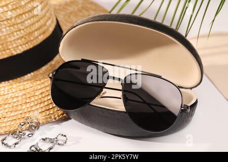 Lunettes de soleil élégantes dans un étui en cuir noir près d'un chapeau de paille et de boucles d'oreilles sur une table blanche Banque D'Images