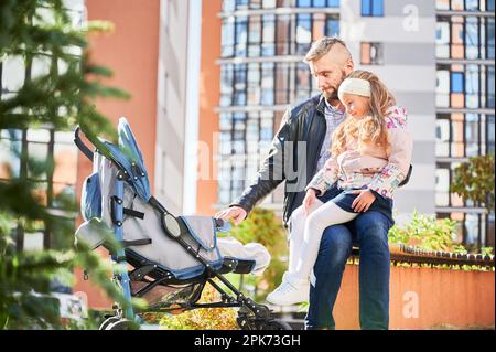 Un père aimant passe du temps avec un nouveau-né à l'extérieur. Vue latérale d'un jeune homme barbu en tenue décontractée marchant avec une poussette dans la rue, avec des immeubles en hauteur en arrière-plan. Concept de parentalité. Banque D'Images