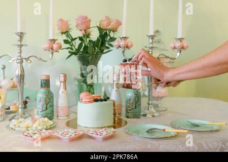 Gros plan du cheesecake de pâques à la main coupant femelle couleur menthe avec des baies fraîches et des macarons, des fleurs, des bonbons sur la table. nature morte avec des roses. Banque D'Images