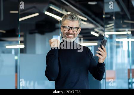 Portrait d'un patron réussi, d'un homme d'affaires célébrant le succès de la victoire et de bons résultats, d'un homme souriant et regardant l'appareil photo, a gagné en ligne au téléphone, employé dans le bureau au travail. Banque D'Images