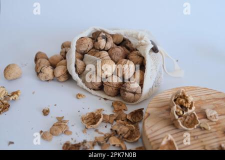 Cerneaux de noyer, coquillages de noyer et un sac en tissu blanc rempli de noix entières sur une table blanche avec une planche à découper. Photo de concept de noix cassées fissurées. Banque D'Images