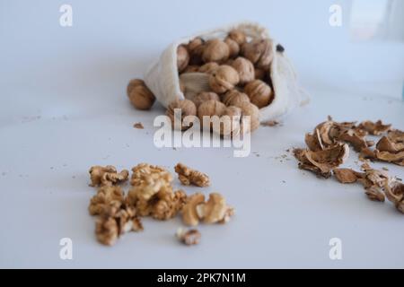 Cerneaux de noyer, coquillages de noyer et un sac en tissu blanc rempli de noix entières sur un fond de table blanc. Idée de photo de concept de noix cassées fissurées. Banque D'Images