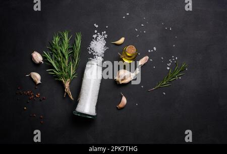 Parsemé de gros sel blanc, de grains de poivre et de brins de romarin sur une table noire, ingrédients pour la cuisson du poisson et de la viande Banque D'Images