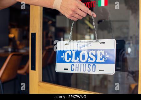 Une personne accrochant une affiche sur une porte de restaurant, lisant Fermer. Fermé, double langue, anglais et italien. Banque D'Images