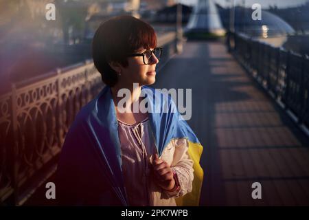 Confiance caucasienne fille en blouse blanche et lunettes couvertes d'un drapeau de l'Ukraine regardant à côté. Portrait de femme patriotique avec drapeau de l'Ukraine au coucher du soleil. Concept de liberté. Photo de haute qualité Banque D'Images