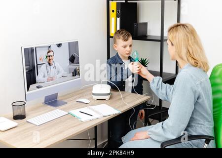 Petit garçon caucasien faisant l'inhalation avec nébuliseur à la maison. Une jeune mère aidant son fils à respirer avec un nébuliseur lors d'une réunion en ligne avec un d Banque D'Images