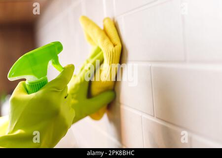 Mains des femmes en gants laver le mur blanc en céramique dans la cuisine avec de la microfibre jaune et un vaporisateur de détergent Banque D'Images