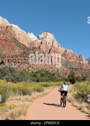 Une jeune femme qui fait du vélo de montagne sur le chemin Pa’rus Trail, un sentier pavé et facile au pied des montagnes abruptes du canyon de Zion, par une journée d'automne claire et ensoleillée Banque D'Images