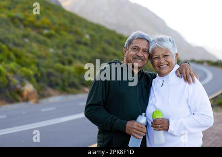 Portrait d'un couple biracial senior heureux portant des vêtements de sport, s'embrassant dans la rue en montagne Banque D'Images