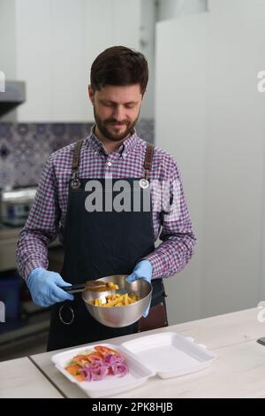 Homme blanc gai préparant une restauration rapide traditionnelle grecque dans une cuisine commerciale. Le cuisinier prépare le plat kalamaki avec des frites et des légumes Banque D'Images