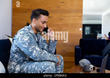 Heureux biracial homme soldat portant un uniforme, assis sur un canapé parlant sur un smartphone dans la salle de séjour. Style de vie, service militaire, communication et gratuit Banque D'Images