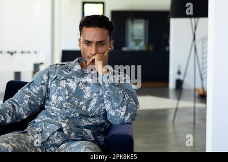 Soldat biracial inquiet portant un uniforme militaire, assis dans un fauteuil dans la salle de séjour à la maison. Style de vie, service militaire et temps libre, unalter Banque D'Images