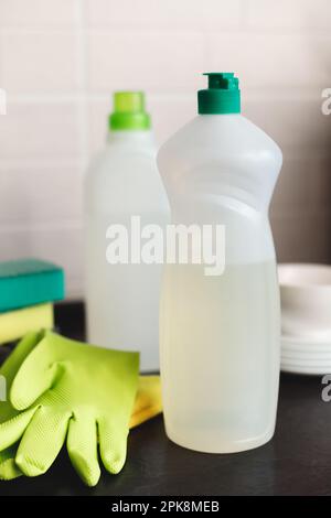 Bouteille en plastique vierge avec détergent liquide pour lave-vaisselle. Gants, éponges et pile de plats sur la table de cuisine. Copier l'espace. Banque D'Images