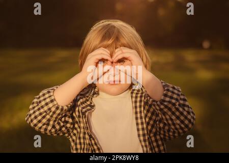 enfant garçon dans un t-shirt blanc décontracté regardant à travers les doigts dans les jumelles gestuelle, observant loin avec un regard attentif, regardant loin. Les mains font du bino Banque D'Images