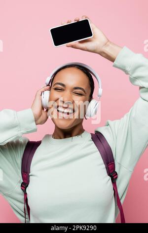 une femme afro-américaine surjoyeuse dans un casque sans fil tenant un smartphone tout en écoutant de la musique isolée sur une image rose Banque D'Images